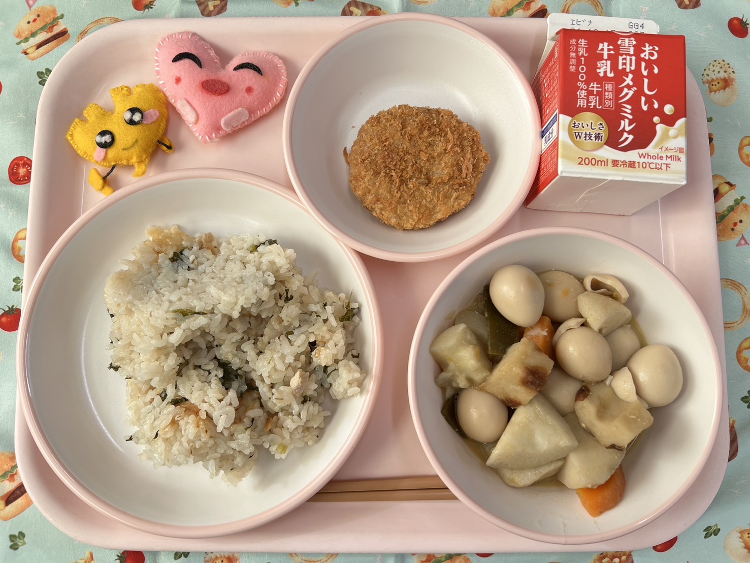 ピンクのトレーに給食が右上から反時計回りで牛乳、メンチカツ、ハートの形をした黄色とピンク色の布製マスコット、とりなめし、おでんと並べられている写真