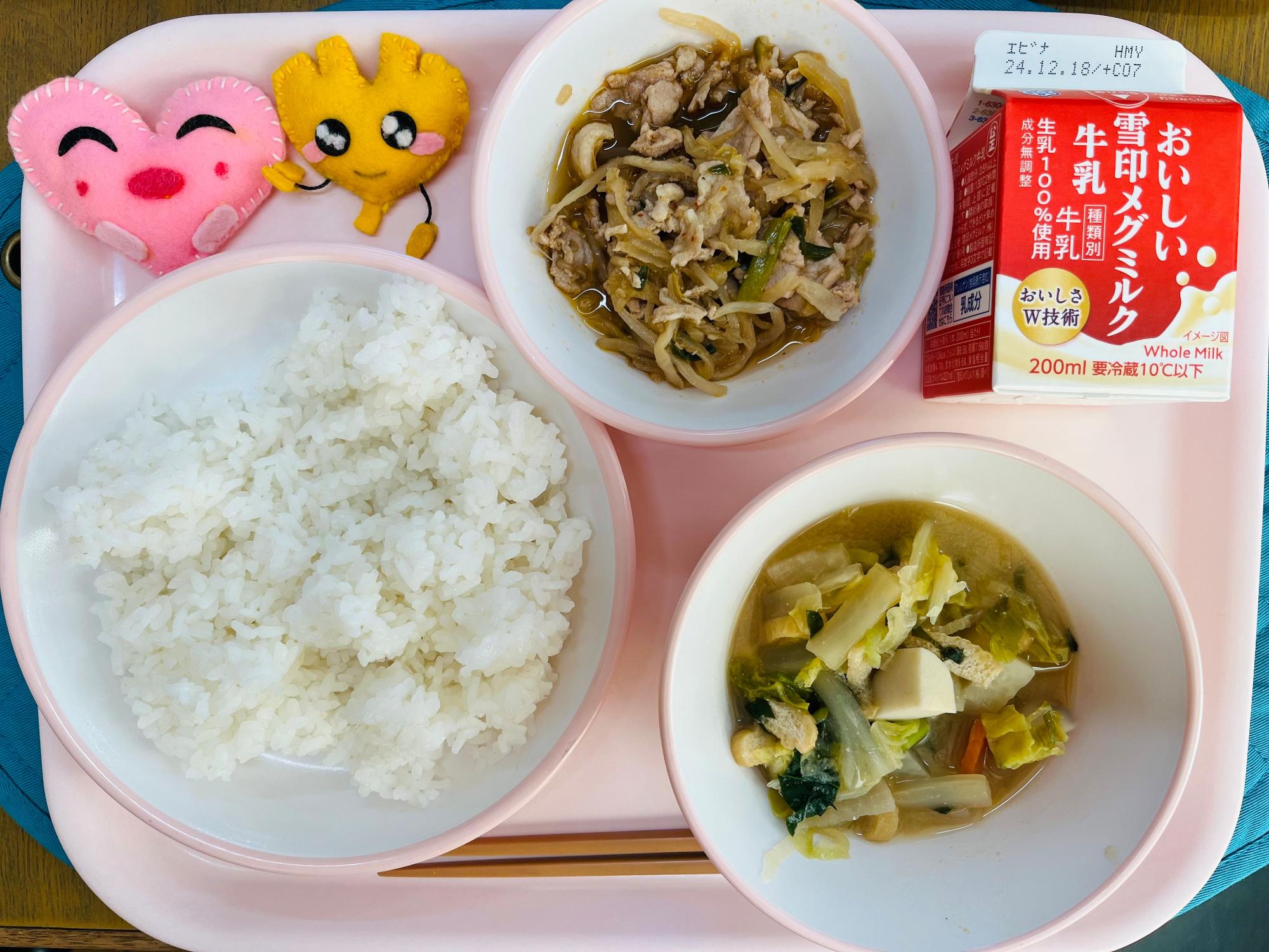 ピンクのトレーに給食が右上から反時計回りで牛乳、豚キムチ、ハートの形をした黄色とピンク色の布製マスコット、ごはん、冬野菜の味噌汁と並べられている写真
