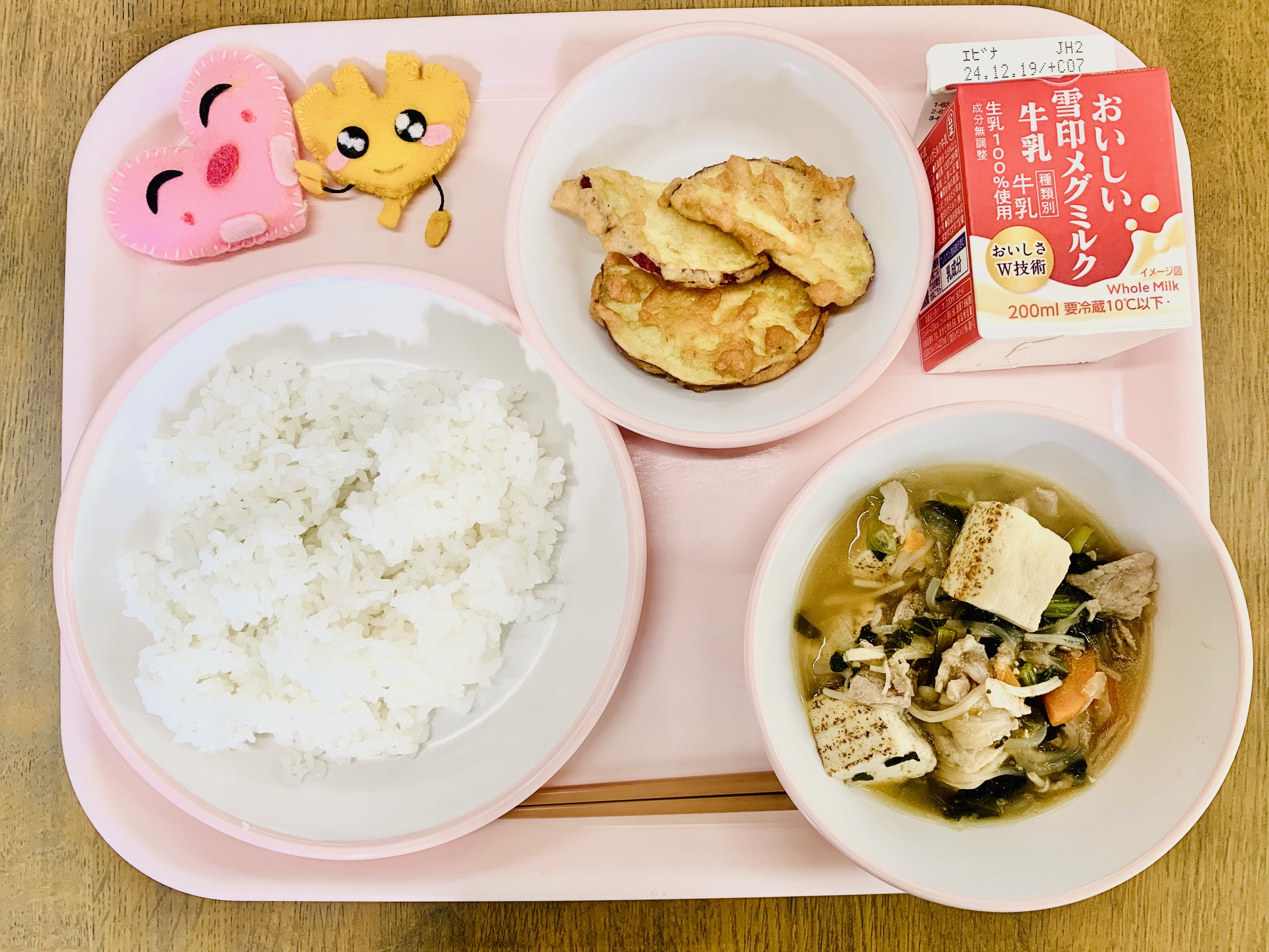 ピンクのトレーに給食が右上から反時計回りで牛乳、サツマイモの天ぷら、ハートの形をした黄色とピンク色の布製マスコット、ごはん、すき焼きと並べられている写真