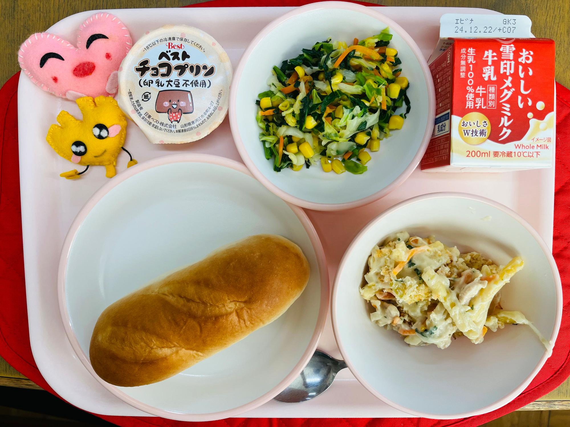 ピンクのトレーに給食が右上から反時計回りで牛乳、野菜ソテー、チョコプリン、ハートの形をした黄色とピンク色の布製マスコット、ミルクパン、マカロニグラタンと並べられている写真