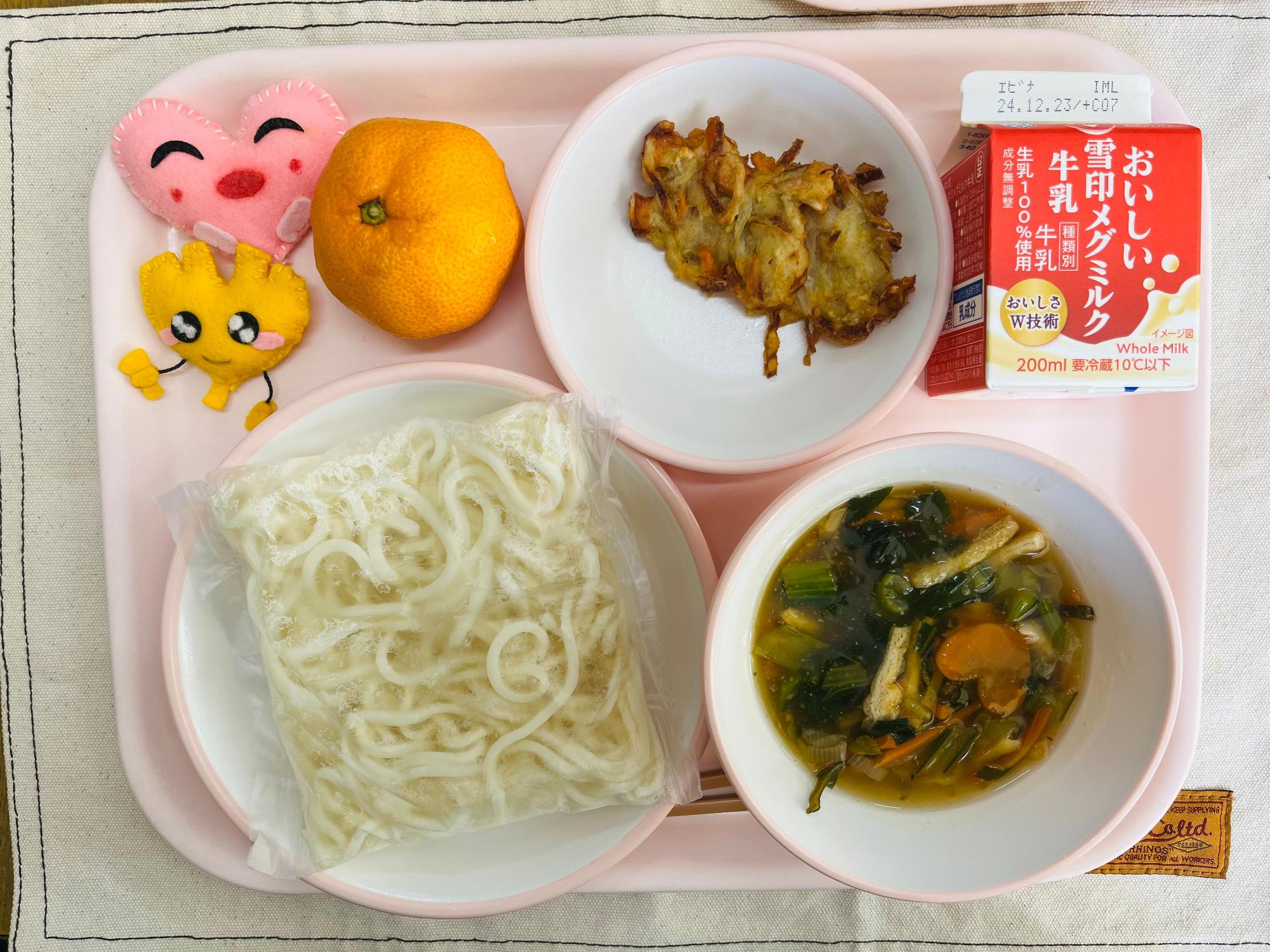 ピンクのトレーに給食が右上から反時計回りで牛乳、ちくわと根菜のかき揚げ、みかん、ハートの形をした黄色とピンク色の布製マスコット、麺、五目スープと並べられている写真