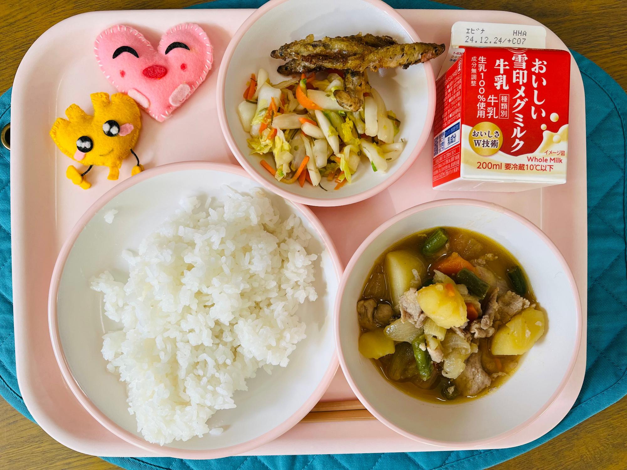 ピンクのトレーに給食が右上から反時計回りで牛乳、白菜漬けとワカサギの唐揚げ、ハートの形をした黄色とピンク色の布製マスコット、ごはん、肉じゃがと並べられている写真