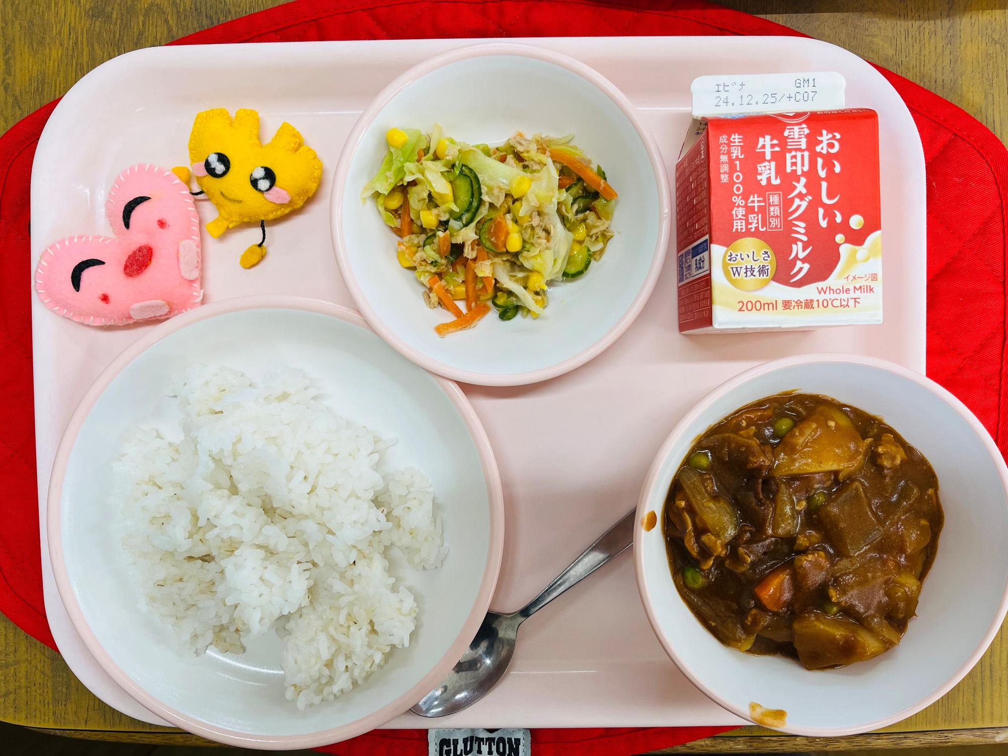 ピンクのトレーに給食が右上から反時計回りで牛乳、ツナコーンサラダ、ハートの形をした黄色とピンク色の布製マスコット、ごはん、ハヤシライスと並べられている写真