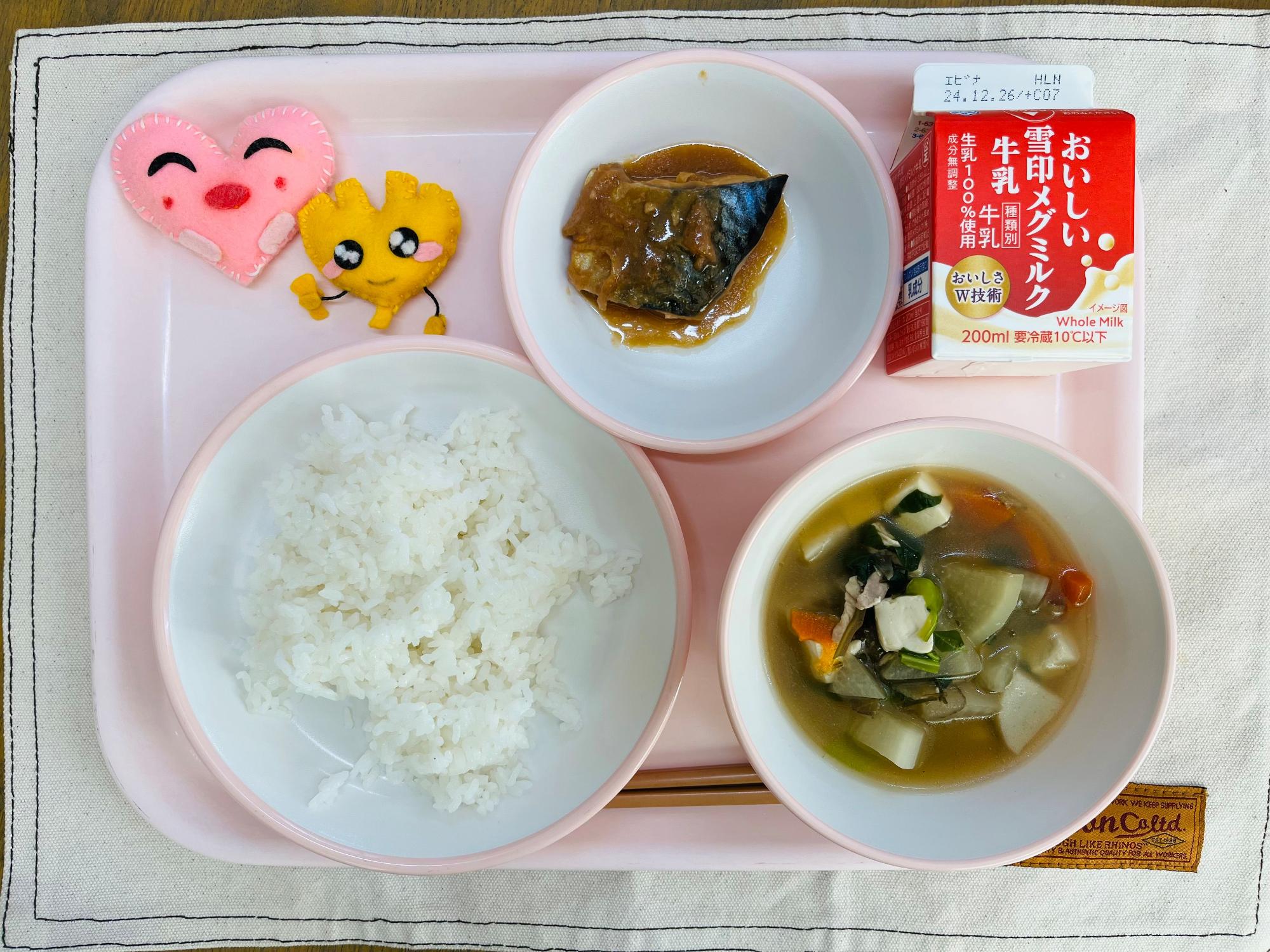 ピンクのトレーに給食が右上から反時計回りで牛乳、サバの味噌煮、ハートの形をした黄色とピンク色の布製マスコット、ごはん、鶏けんちん汁と並べられている写真