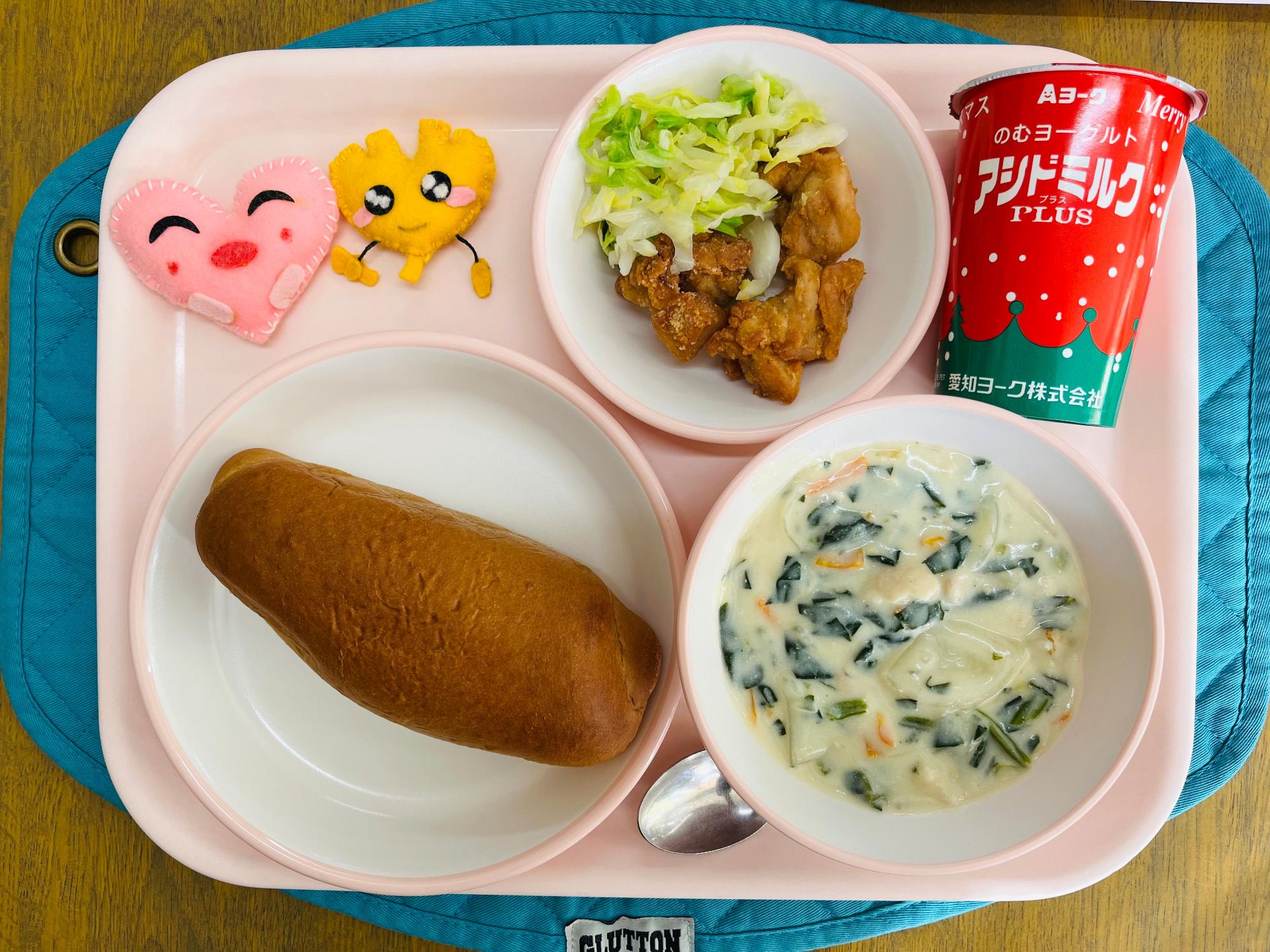 ピンクのトレーに給食が右上から反時計回りで発酵乳、キャベツソテーと鶏肉のからあげ、ハートの形をした黄色とピンク色の布製マスコット、くろパン、ほうれん草のミルクスープと並べられている写真