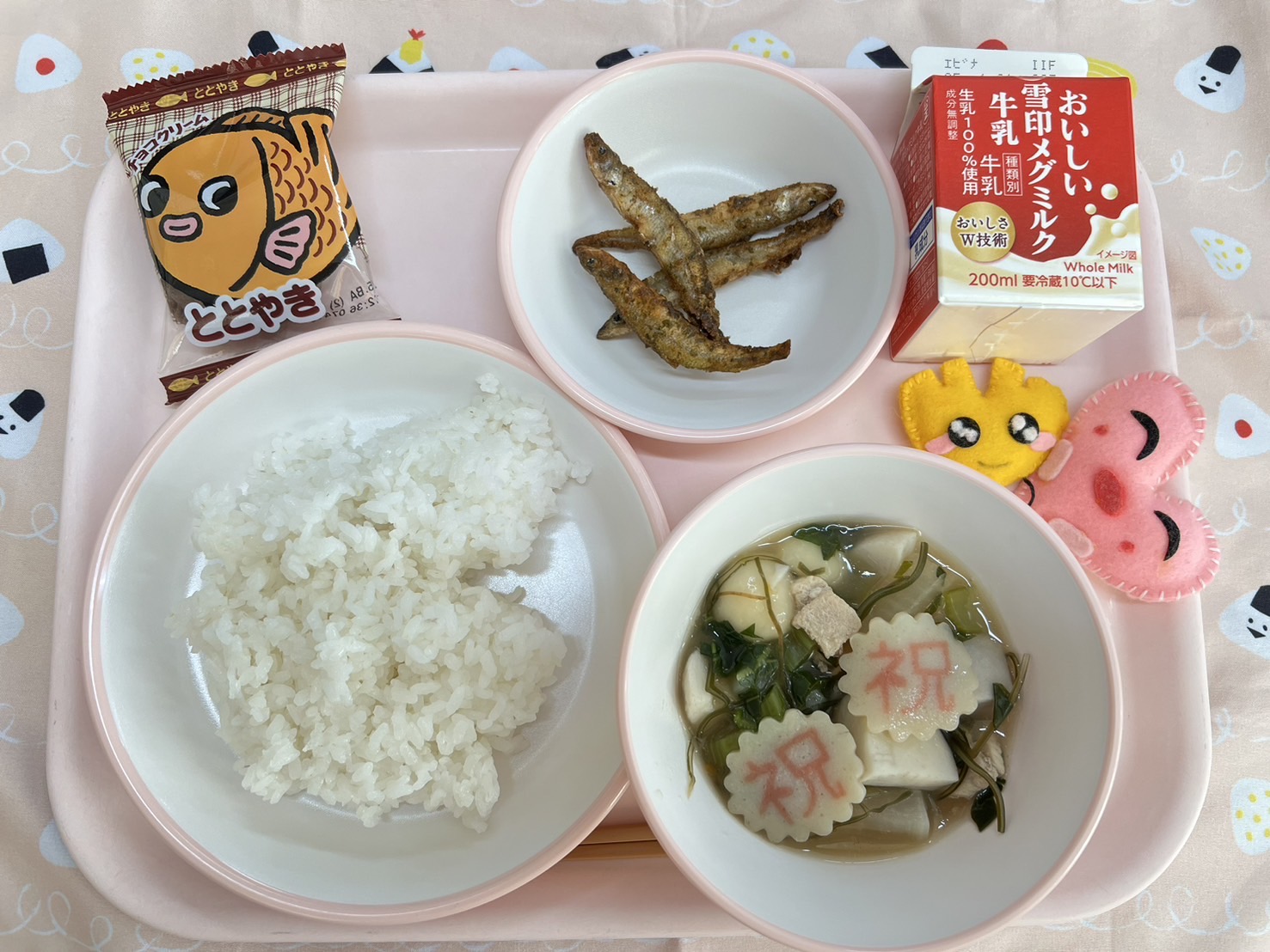 ピンクのトレーに給食が右上から反時計回りで牛乳、わかさぎの唐揚げ、ととやき、ハートの形をした黄色とピンク色の布製マスコット、ごはん、白玉雑煮と並べられている写真