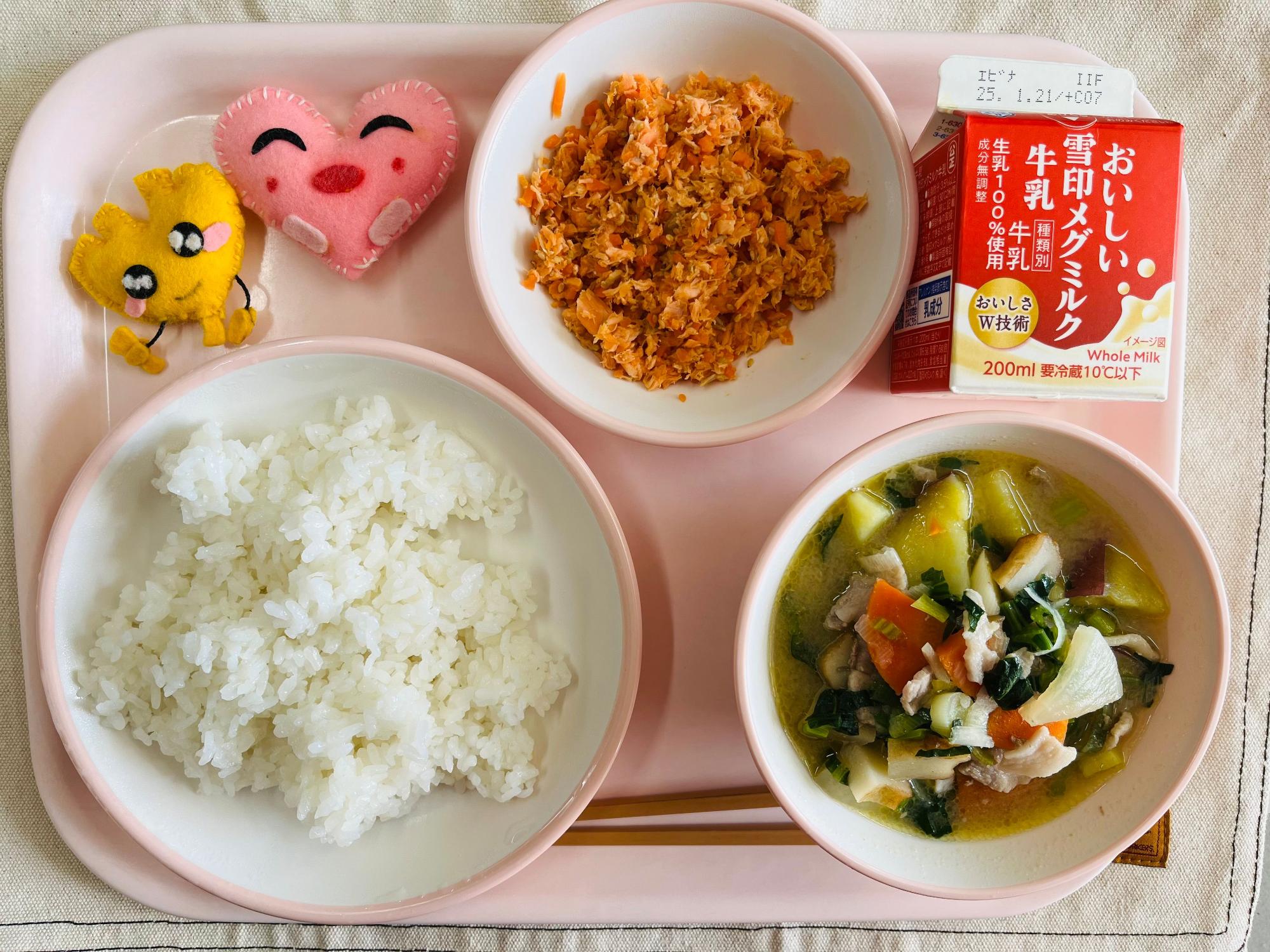 ピンクのトレーに給食が右上から反時計回りで牛乳、さけそぼろ、ハートの形をした黄色とピンク色の布製マスコット、ごはん、さつま汁と並べられている写真