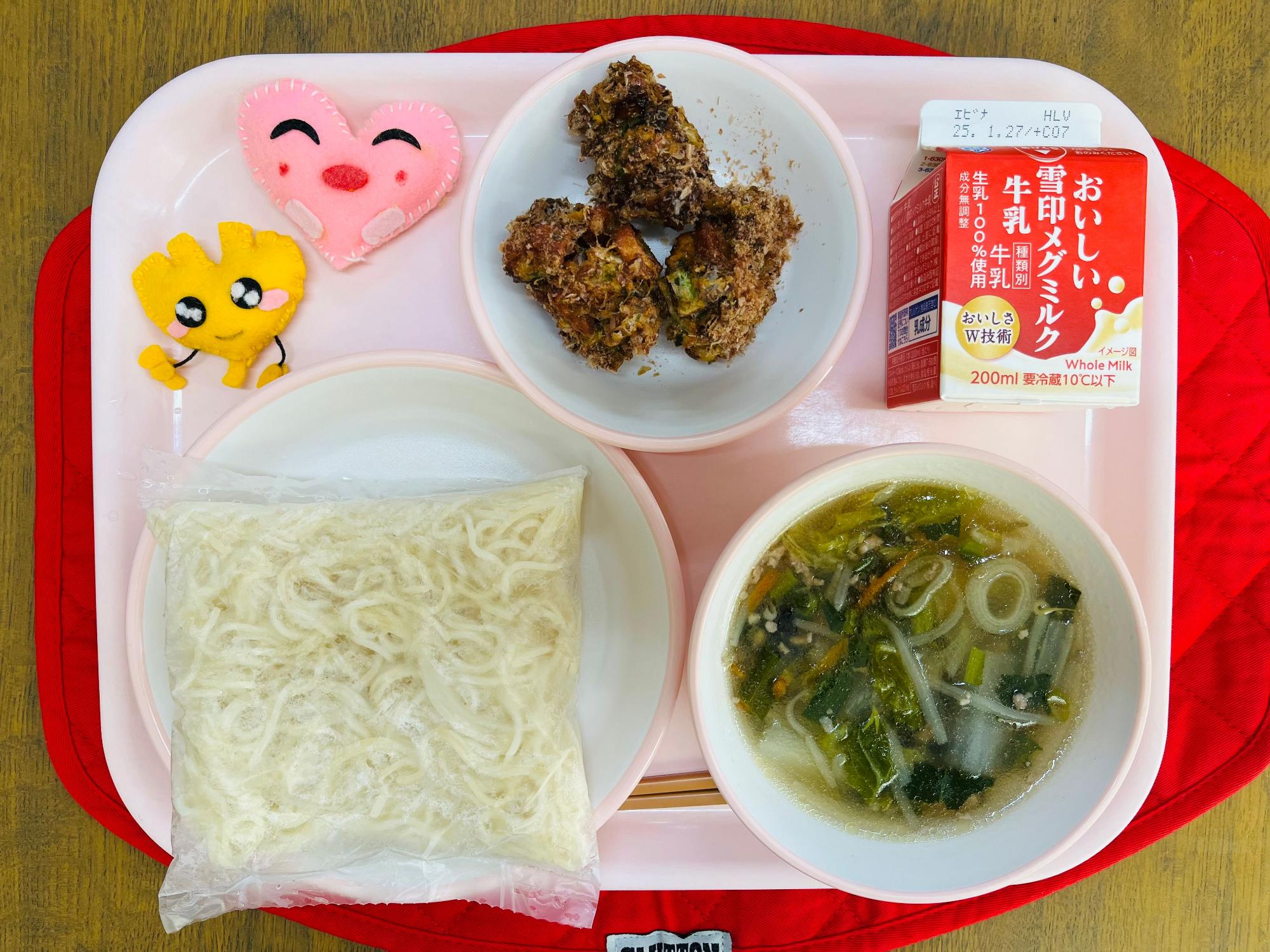 ピンクのトレーに給食が右上から反時計回りで牛乳、お好みちくわボール、ハートの形をした黄色とピンク色の布製マスコット、麺、塩ラーメンのスープと具が並べられている写真