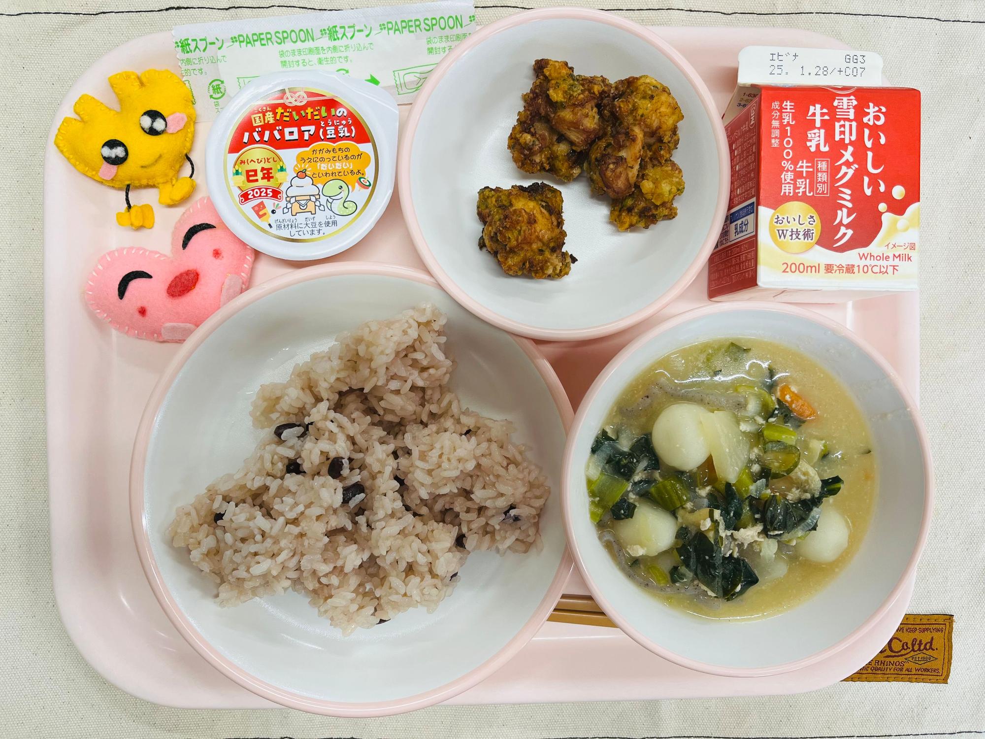 ピンクのトレーに給食が右上から反時計回りで牛乳、鶏肉のふうみあげ、だいだいのババロア、ハートの形をした黄色とピンク色の布製マスコット、あずきごはん、だんご汁と並べられている写真