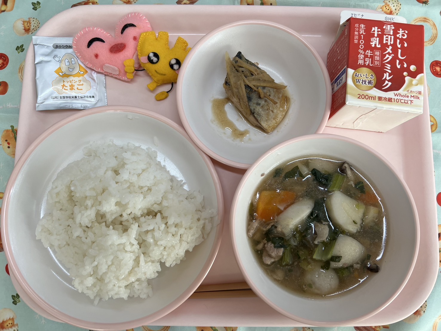 ピンクのトレーに給食が右上から反時計回りで牛乳、鯖の生姜煮、ハートの形をした黄色とピンク色の布製マスコット、たまごふりかけ、ごはん、のっぺい汁と並べられている写真