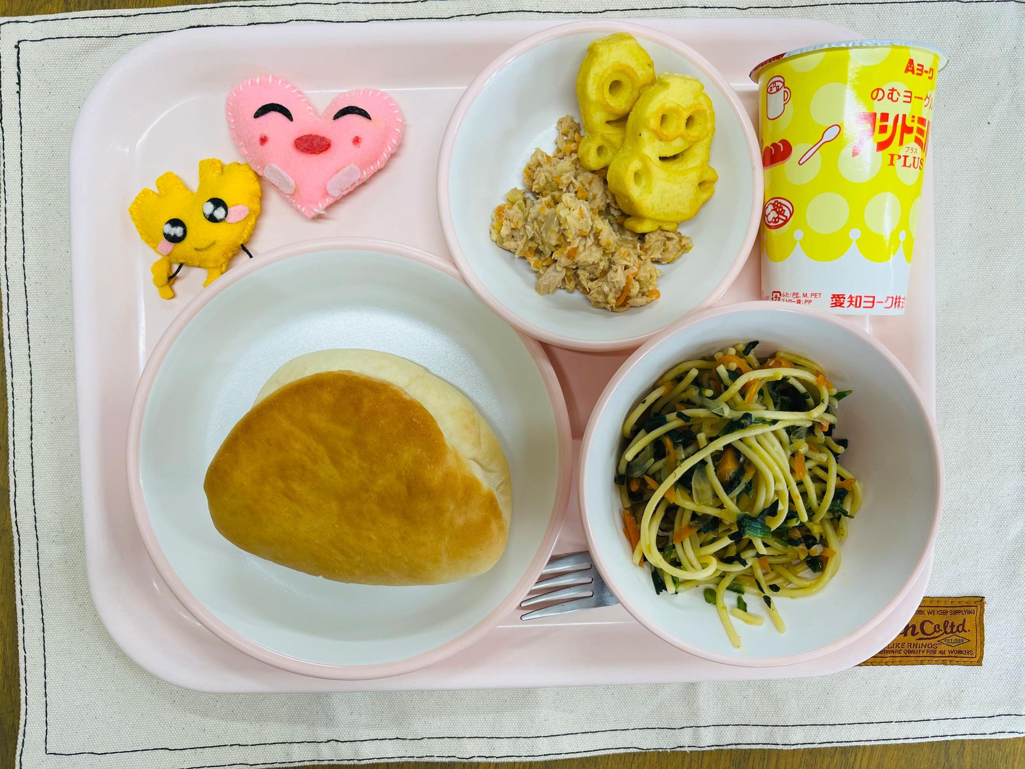 ピンクのトレーに給食が右上から反時計回りで発酵乳、おたのしみポテトとツナサンドの具、ハートの形をした黄色とピンク色の布製マスコット、サンドパン、ほうれん草とベーコンのスパゲッティと並べられている写真