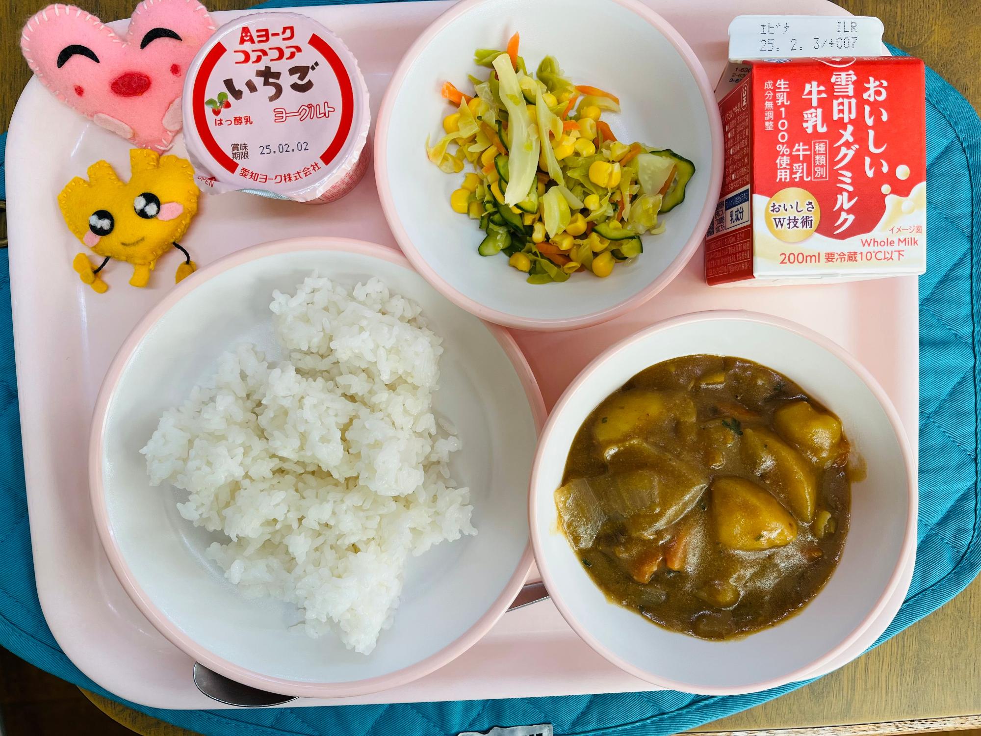 ピンクのトレーに給食が右上から反時計回りで牛乳、コーンサラダ、ヨーグルト、ハートの形をした黄色とピンク色の布製マスコット、ごはん、チキンカレーと並べられている写真