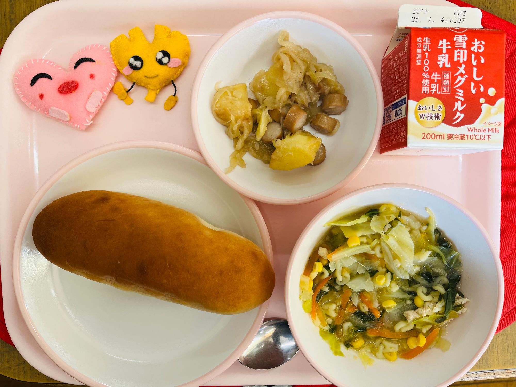 ピンクのトレーに給食が右上から反時計回りで牛乳、ジャーマンポテト、ハートの形をした黄色とピンク色の布製マスコット、あじつけパン、ABCスープと並べられている写真