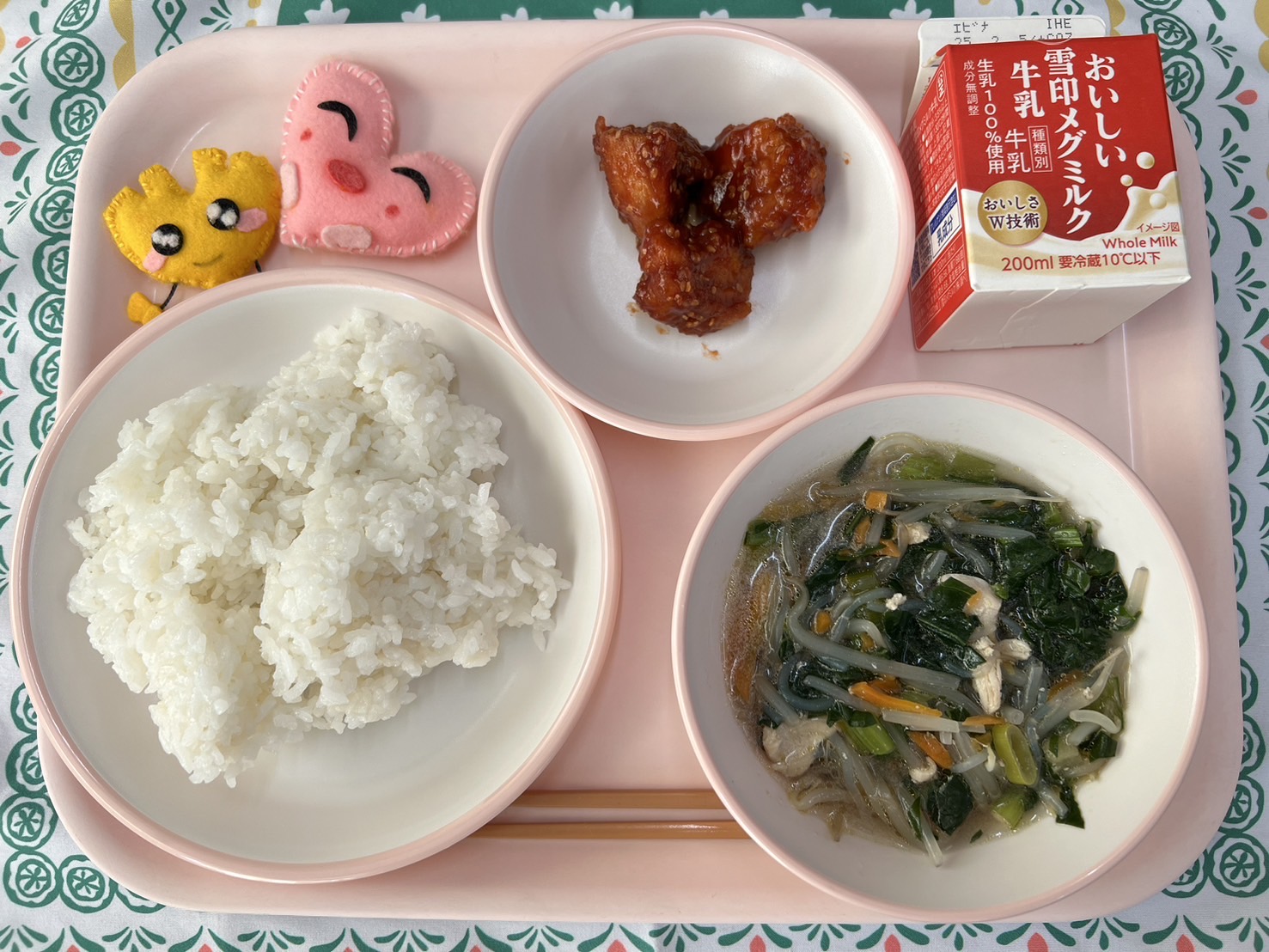 ピンクのトレーに給食が右上から反時計回りで牛乳、ヤンニョムチキン、ハートの形をした黄色とピンク色の布製マスコット、ごはん、五目スープと並べられている写真