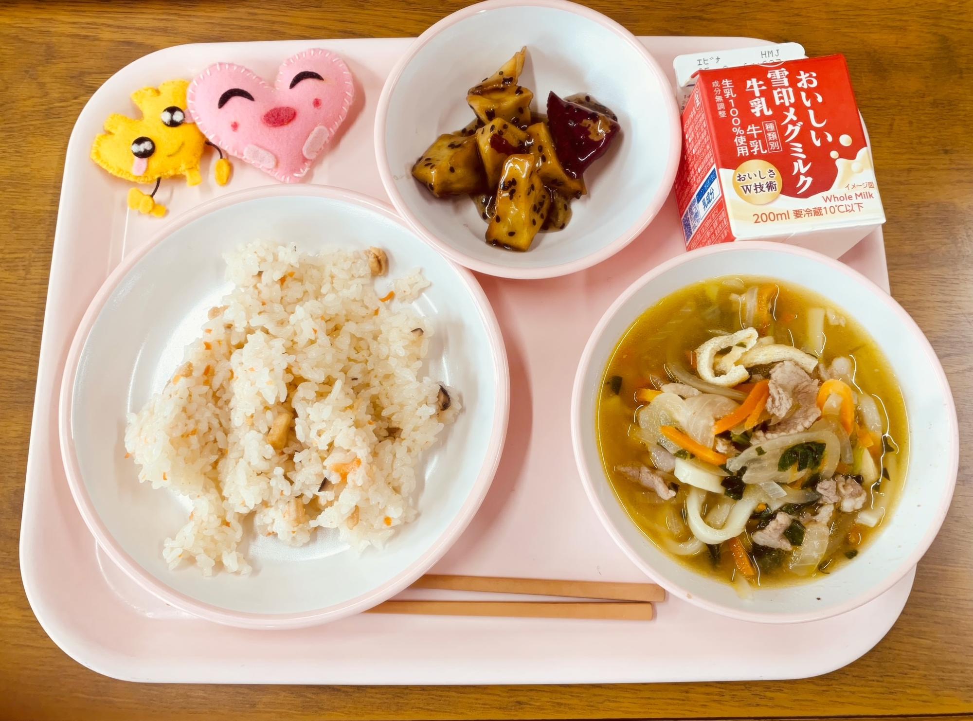 ピンクのトレーに給食が右上から反時計回りで牛乳、大学芋、ハートの形をした黄色とピンク色の布製マスコット、かしわずし、肉うどんと並べられている写