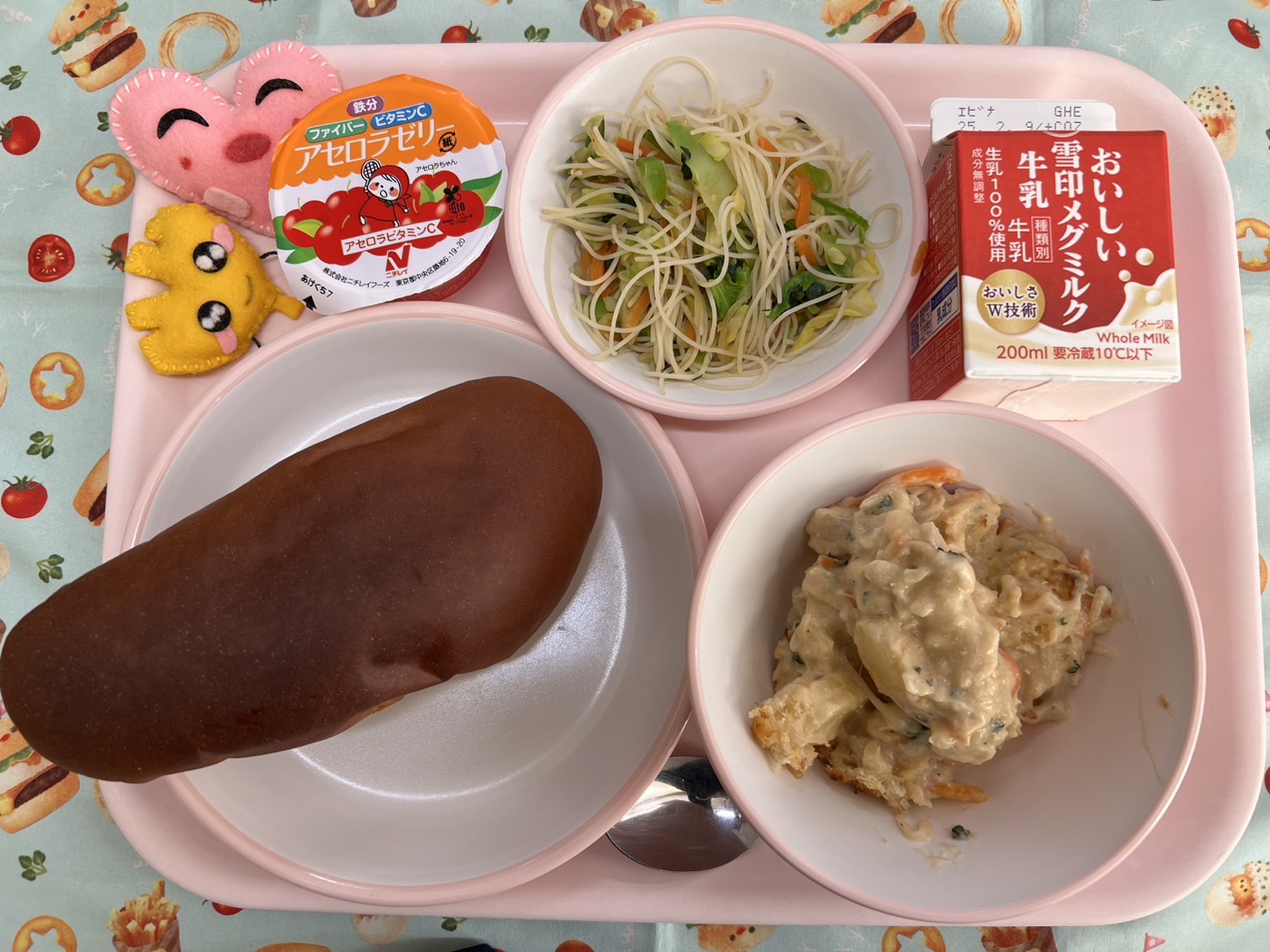 ピンクのトレーに給食が右上から反時計回りで牛乳、ビーフンソテー、アセロラゼリー、ハートの形をした黄色とピンク色の布製マスコット、くろパン、ポテトグラタンと並べられている写真