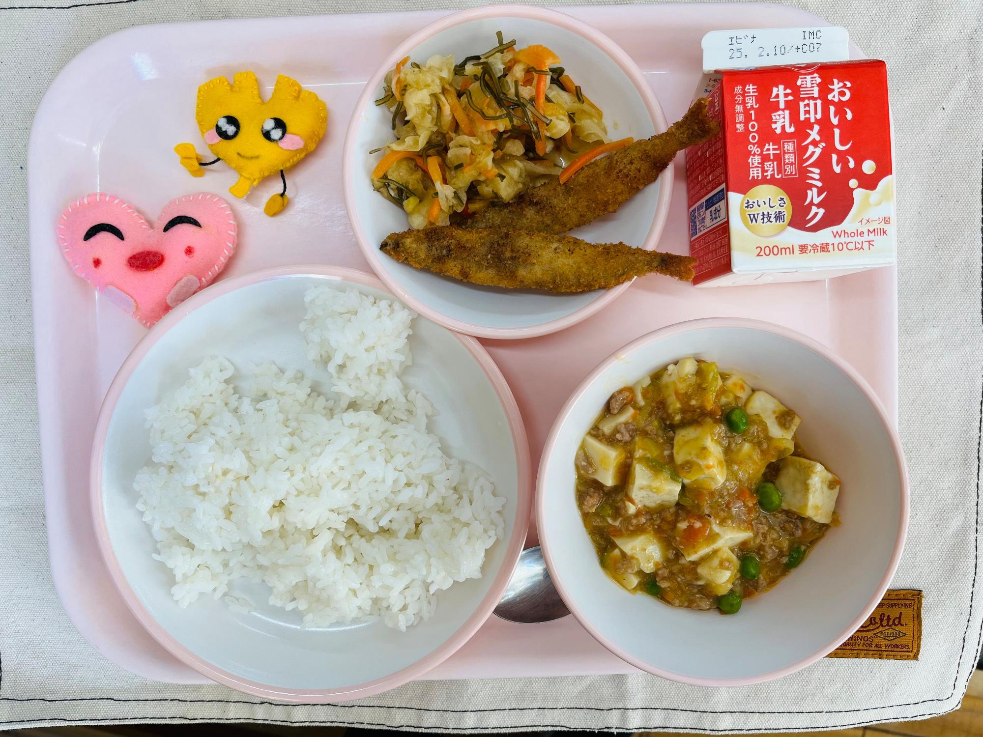 ピンクのトレーに給食が右上から反時計回りで牛乳、ピリ辛大根とししゃもフライ、ハートの形をした黄色とピンク色の布製マスコット、ごはん、マーボー豆腐と並べられている写真