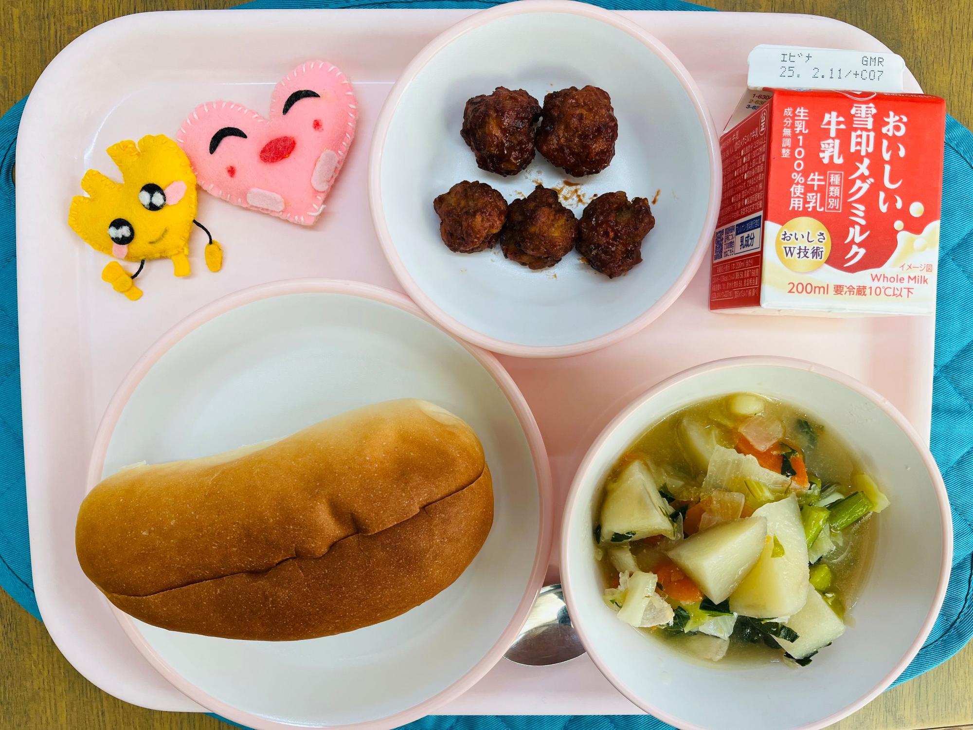 ピンクのトレーに給食が右上から反時計回りで牛乳、ミートボール、ハートの形をした黄色とピンク色の布製マスコット、サンドパン、野菜のスープ煮と並べられている写真