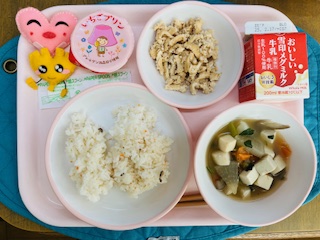 ピンクのトレーに給食が右上から反時計回りで牛乳、こぎつね寿司の具、いちごプリン、ハートの形をした黄色とピンク色の布製マスコット、酢飯、けんちん汁と並べられている写真