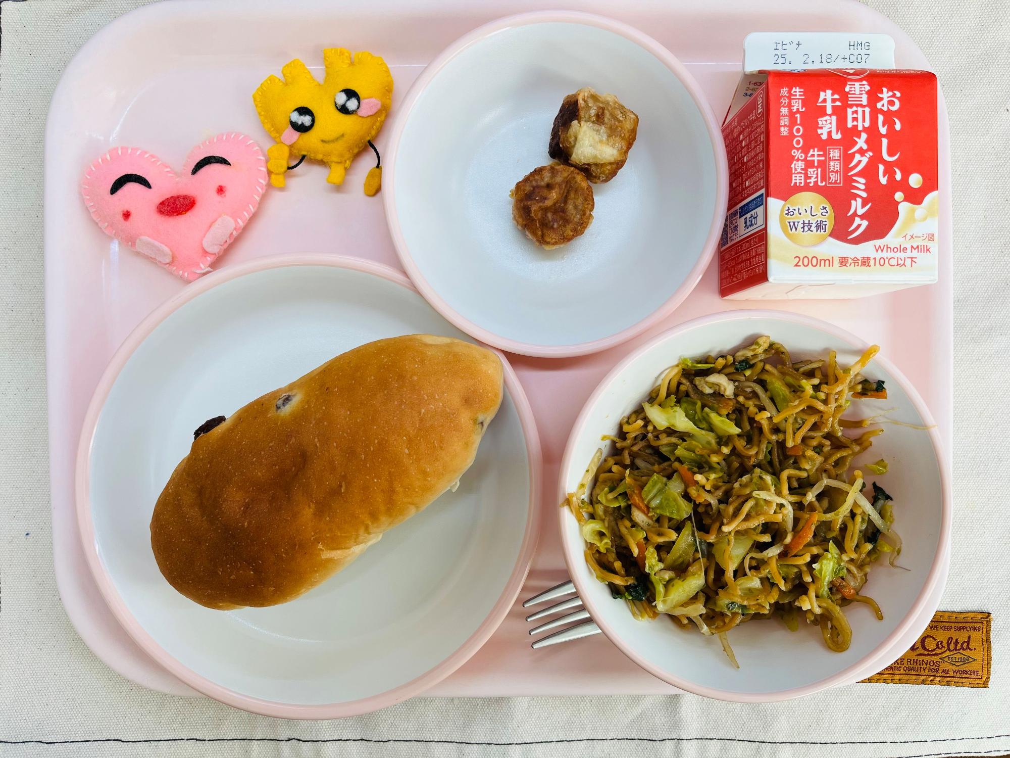 ピンクのトレーに給食が右上から反時計回りで牛乳、揚げシュウマイ、ハートの形をした黄色とピンク色の布製マスコット、ぶどうパン、ソース焼きそばと並べられている写真
