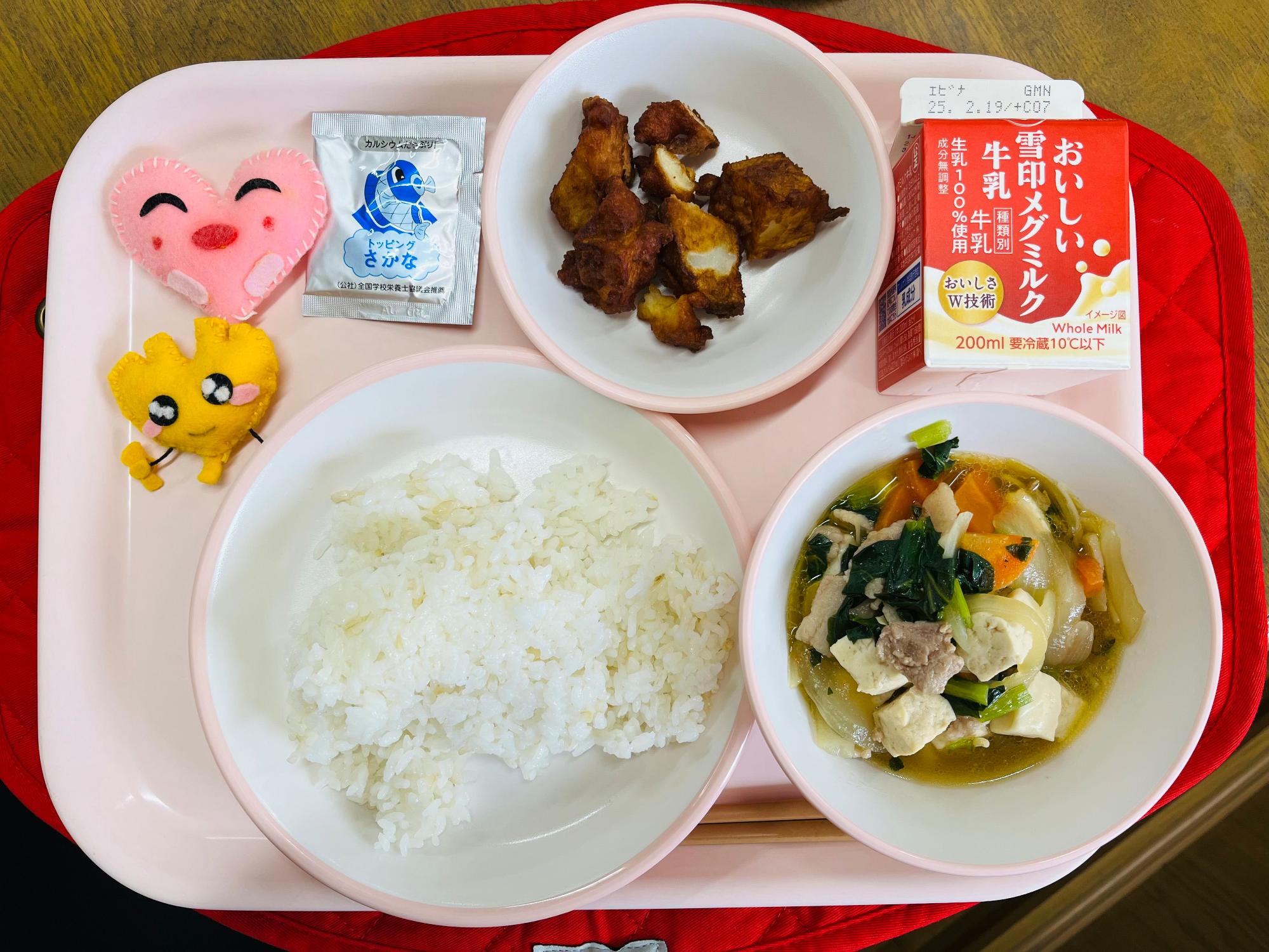 ピンクのトレーに給食が右上から反時計回りで牛乳、じゃがじゃが、魚ふりかけ、ハートの形をした黄色とピンク色の布製マスコット、発芽玄米ごはん、肉豆腐と並べられている写真