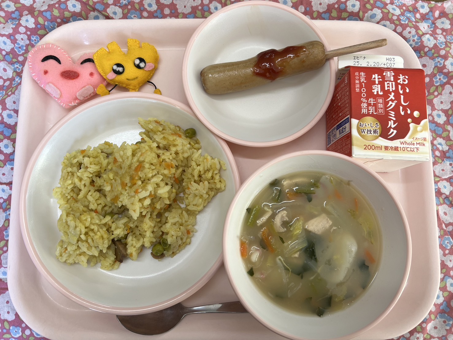 ピンクのトレーに給食が右上から反時計回りで牛乳、フランクフルト、ハートの形をした黄色とピンク色の布製マスコット、ベジカレーピラフ、トックスープと並べられている写真