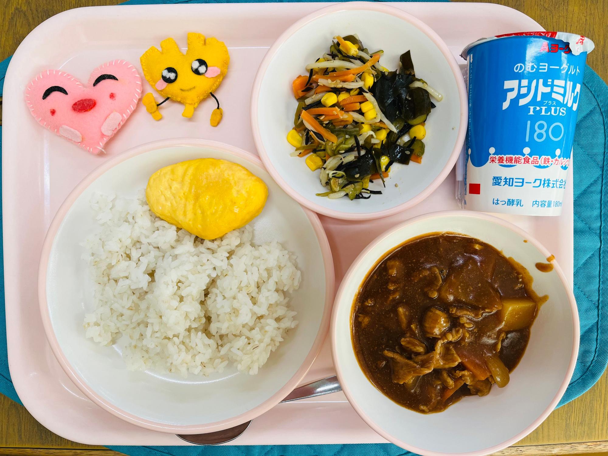 ピンクのトレーに給食が右上から反時計回りで発酵乳、海藻サラダ、ハートの形をした黄色とピンク色の布製マスコット、オムレツとごはん、ハヤシライスと並べられている写真