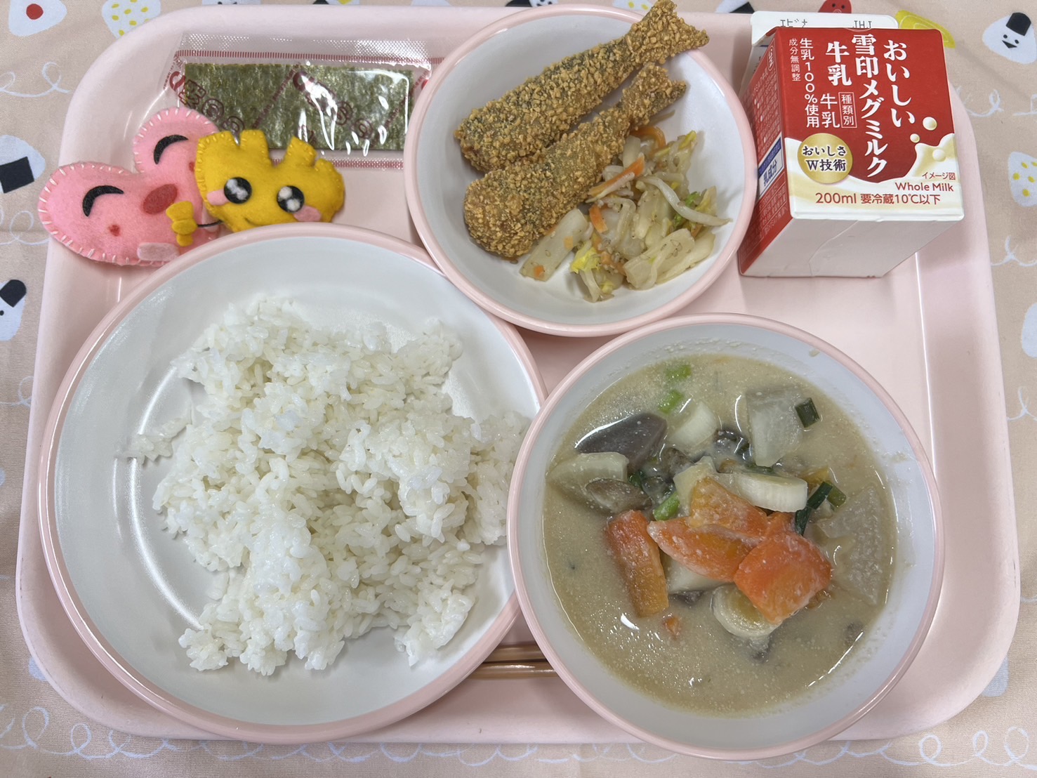 ピンクのトレーに給食が右上から反時計回りで牛乳、野菜の胡麻和えといわしのカリカリ揚げ、焼き海苔、ハートの形をした黄色とピンク色の布製マスコット、ごはん、ごじると並べられている写真
