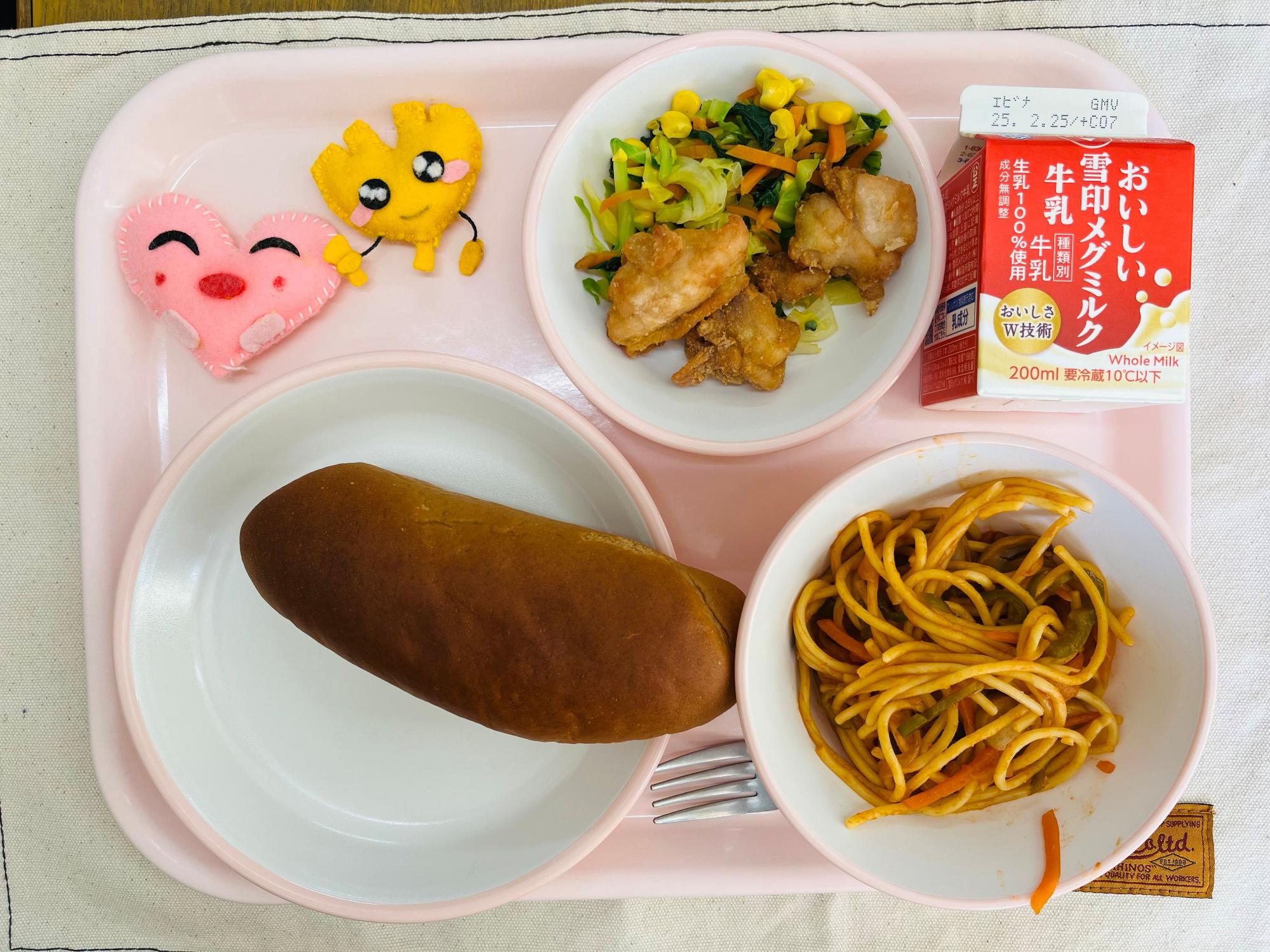 ピンクのトレーに給食が右上から反時計回りで牛乳、野菜ソテーと鶏肉のからあげ、ハートの形をした黄色とピンク色の布製マスコット、くろパン、しろくまスパゲッティと並べられている写真