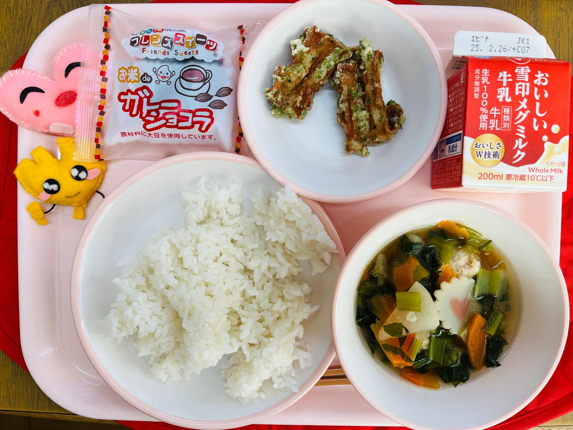 ピンクのトレーに給食が右上から反時計回りで牛乳、ちくわの磯辺揚げ、ガトーショコラ、ハートの形をした黄色とピンク色の布製マスコット、ごはん、大根と肉団子のスープと並べられている写真