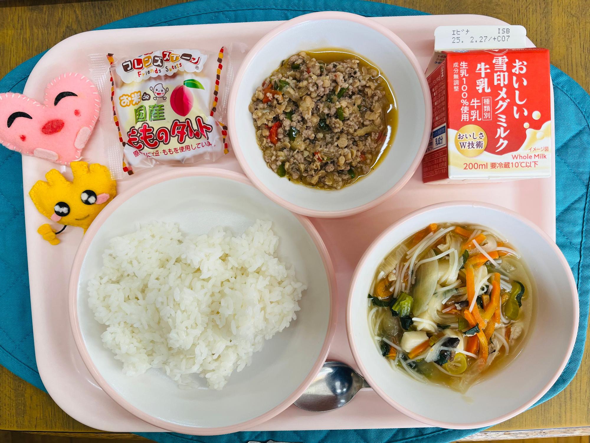 ピンクのトレーに給食が右上から反時計回りで牛乳、ガパオライスの具、ももタルト、ハートの形をした黄色とピンク色の布製マスコット、ごはん、ビーフンスープと並べられている写真
