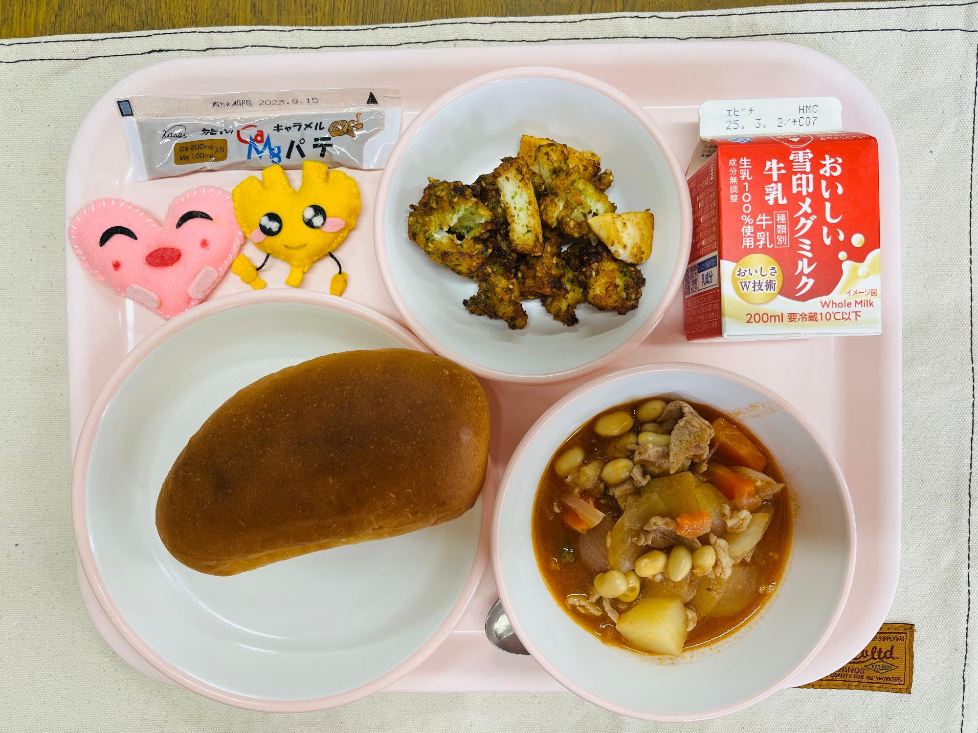 ピンクのトレーに給食が右上から反時計回りで牛乳、いかの風味揚げ、キャラメルパテ、ハートの形をした黄色とピンク色の布製マスコット、味付けパン、ポークビーンズと並べられている写真