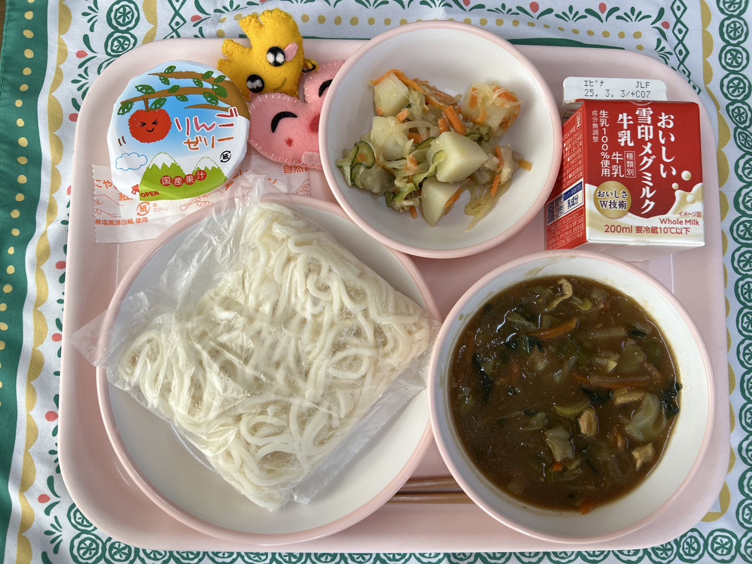 ピンクのトレーに給食が右上から反時計回りで牛乳、ジャーマンポテト、ハートの形をした黄色とピンク色の布製マスコット、りんごゼリー、ソフト麺、カレーあんかけと並べられている写真