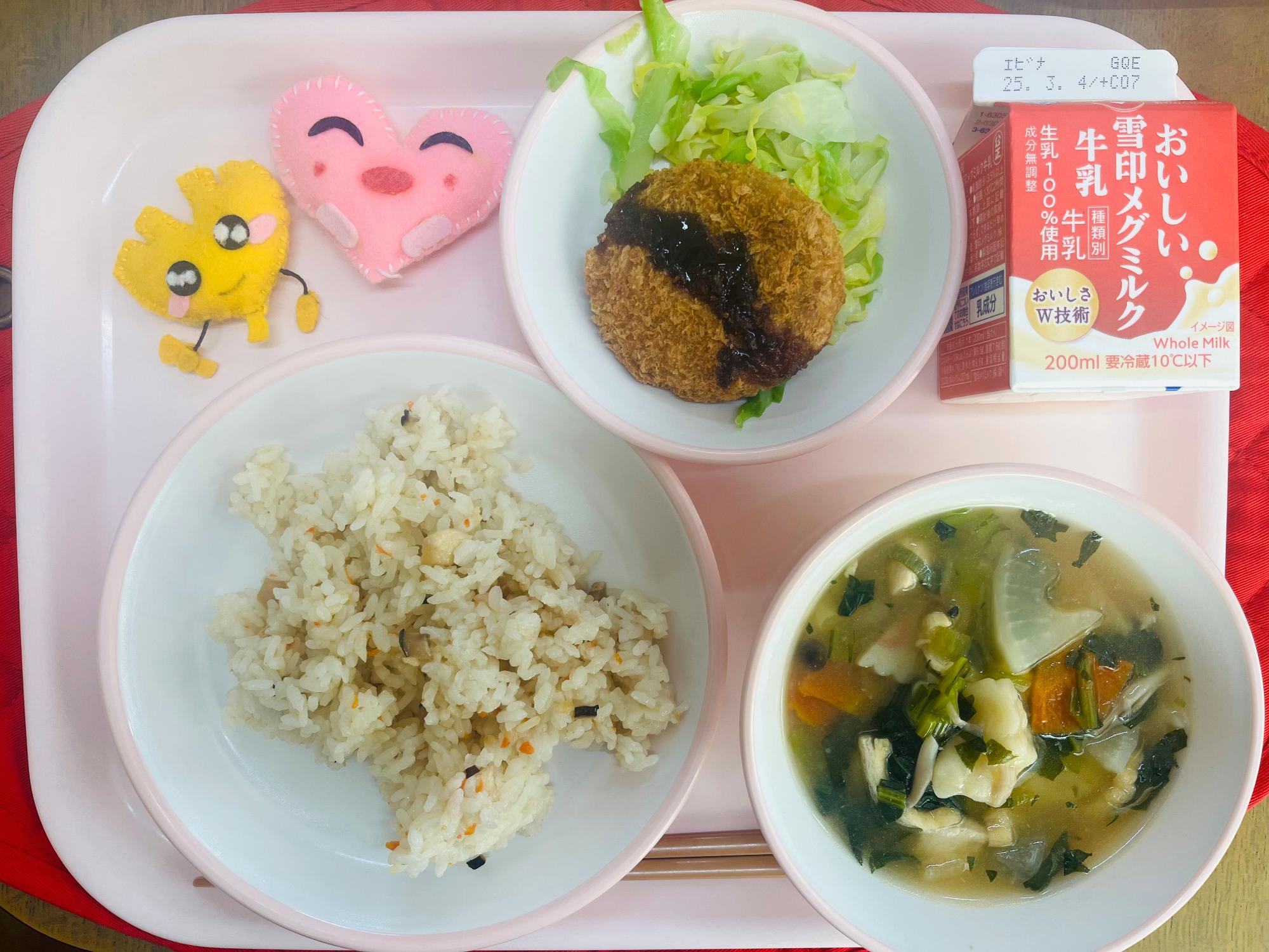 ピンクのトレーに給食が右上から反時計回りで牛乳、キャベツソテーとたまねぎメンチカツ、ハートの形をした黄色とピンク色の布製マスコット、かやくごはん、すいとんと並べられている写真