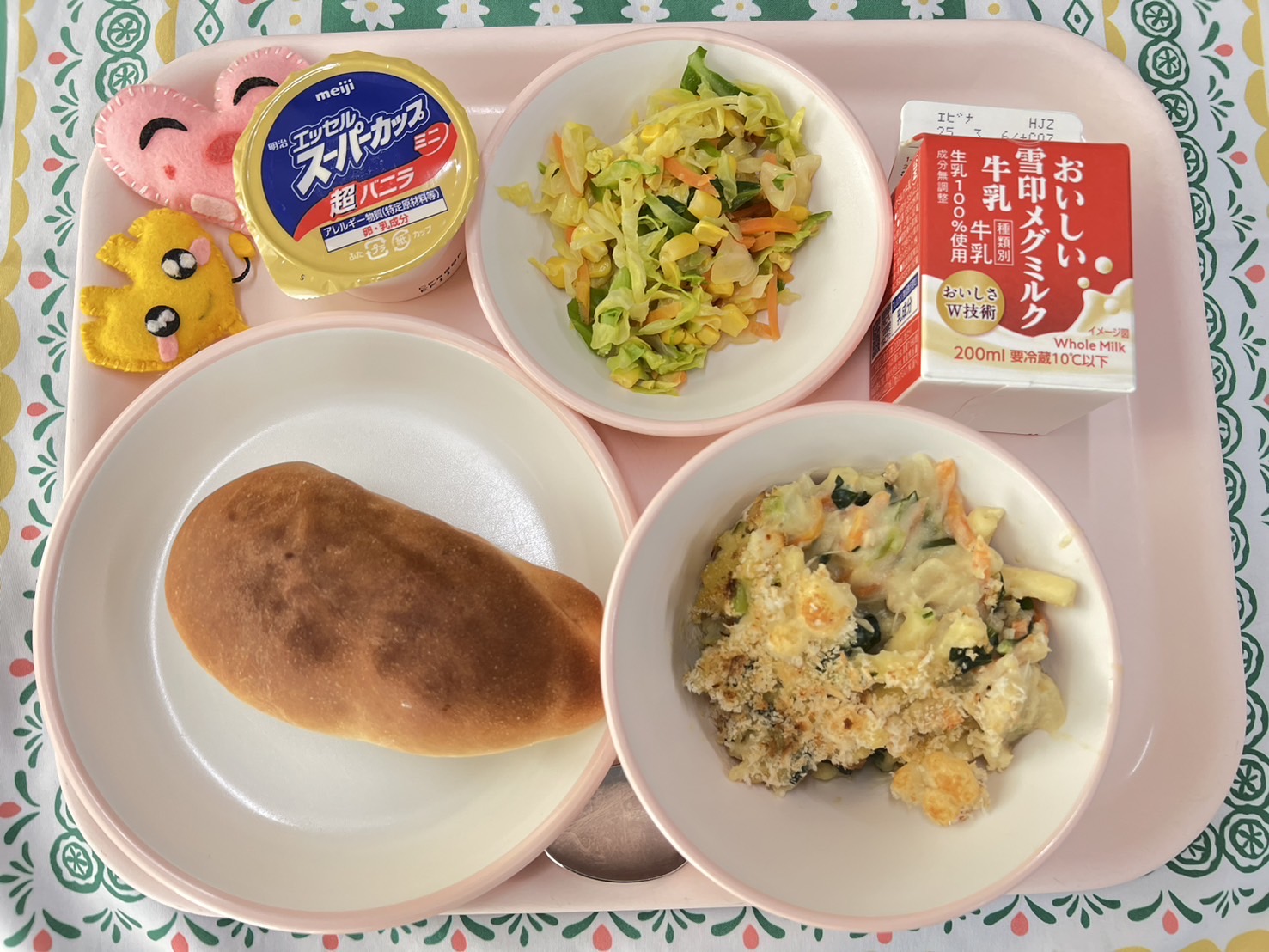 ピンクのトレーに給食が右上から反時計回りで牛乳、ミックスソテー、バニラアイス、ハートの形をした黄色とピンク色の布製マスコット、ソフトフランスパン、マカロニの豆乳グラタンと並べられている写真