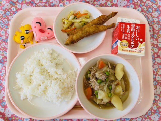 ピンクのトレーに給食が右上から反時計回りで牛乳、白菜漬けとししゃもフライ、ハートの形をした黄色とピンク色の布製マスコット、ごはん、肉じゃがと並べられている写真