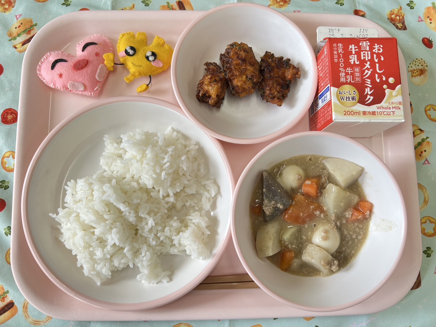 ピンクのトレーに給食が右上から反時計回りで牛乳、さわらのころも揚げ、ハートの形をした黄色とピンク色の布製マスコット、ごはん、肉味噌おでんと並べられている写真