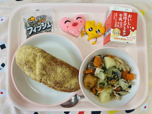 ピンクのトレーに給食が右上から反時計回りで牛乳、ハートの形をした黄色とピンク色の布製マスコット、味付け小魚、きなこ揚げパン、和風ポトフと並べられている写真