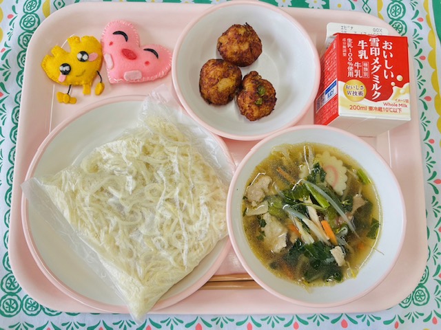 ピンクのトレーに給食が右上から反時計回りで牛乳、じゃがいもの一口揚げ、ハートの形をした黄色とピンク色の布製マスコット、麺、しょうゆラーメンの具とスープが並べられている写真