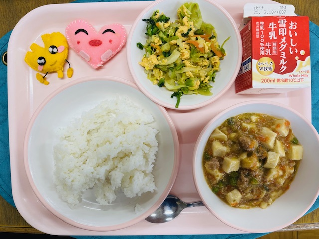 ピンクのトレーに給食が右上から反時計回りで牛乳、たまご和え、ハートの形をした黄色とピンク色の布製マスコット、ごはん、マーボー豆腐と並べられている写真