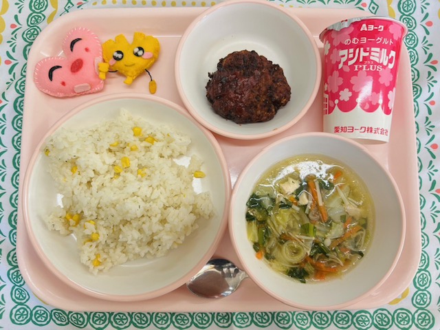 ピンクのトレーに給食が右上から反時計回りで発酵乳、ビーンズハンバーグ、ハートの形をした黄色とピンク色の布製マスコット、コーンピラフ、コンソメスープと並べられている写真