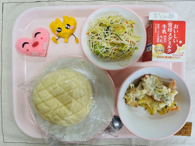 ピンクのトレーに給食が右上から反時計回りで牛乳、ビーフンソテー、ハートの形をした黄色とピンク色の布製マスコット、メロンパン、マカロニグラタンと並べられている写真