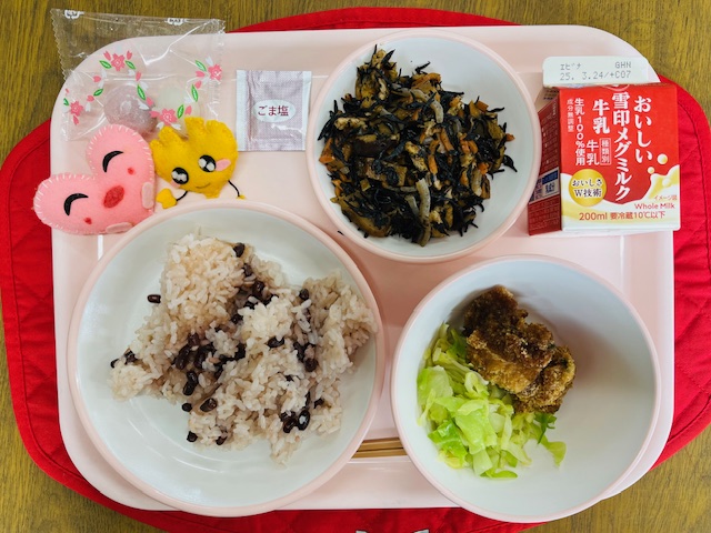 ピンクのトレーに給食が右上から反時計回りで牛乳、ひじきの炒り煮、ごましお、紅白大福、ハートの形をした黄色とピンク色の布製マスコット、赤飯、キャベツソテー、チキンカツと並べられている写真