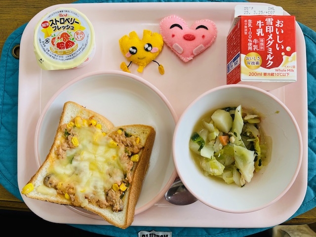 ピンクのトレーに給食が右上から反時計回りで牛乳、ハートの形をした黄色とピンク色の布製マスコット、いちごゼリー、ツナピザトースト、野菜のスープ煮と並べられている写真