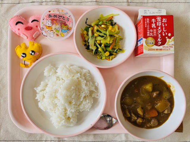 ピンクのトレーに給食が右上から反時計回りで牛乳、野菜ソテー 、おめでとうゼリー、ハートの形をした黄色とピンク色の布製マスコット、ごはん、カレールーと並べられている写真