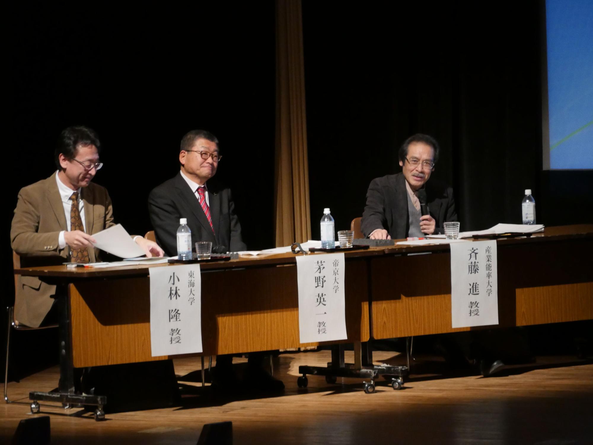 照明を落とした会場でスポットライトの下のテーブルに3人の男性が座っており、右側の男性がマイクを持って発言している様子の写真