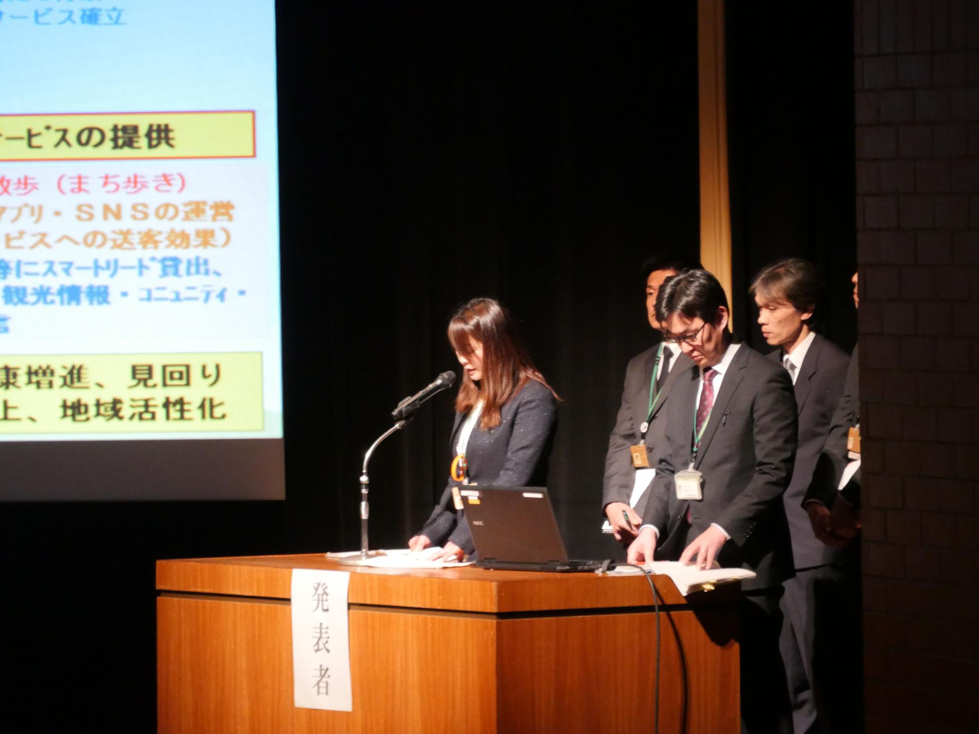 スクリーンの前の演台に男性と女性が並んで立ち、後ろに数人のスーツ姿の男性が立っている発表の様子の写真