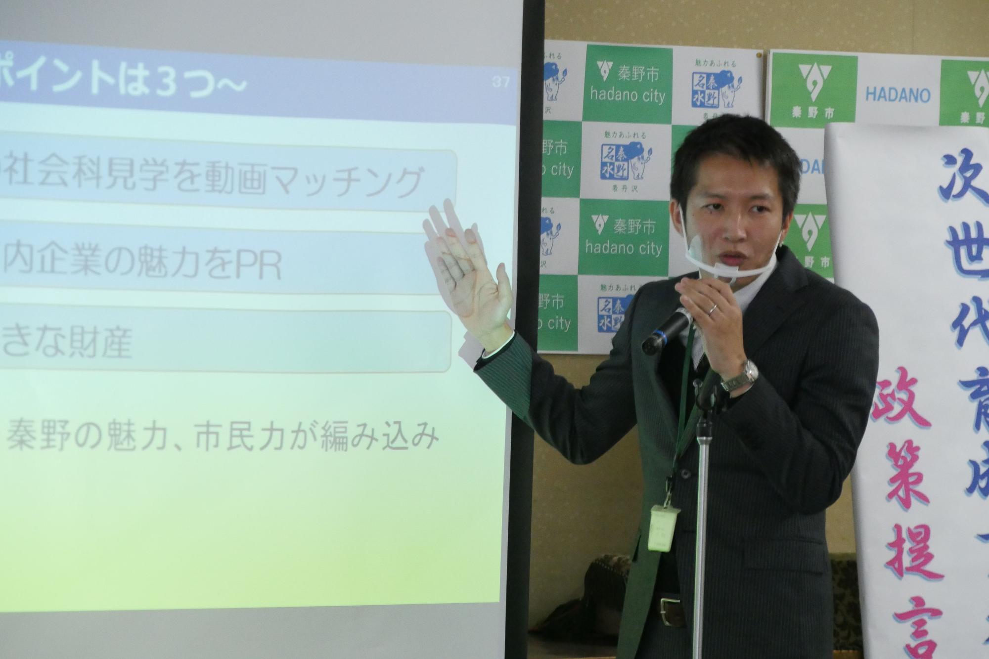 男性がマイクを持って立ち、左側のスクリーンに映し出された資料を説明している場面の写真