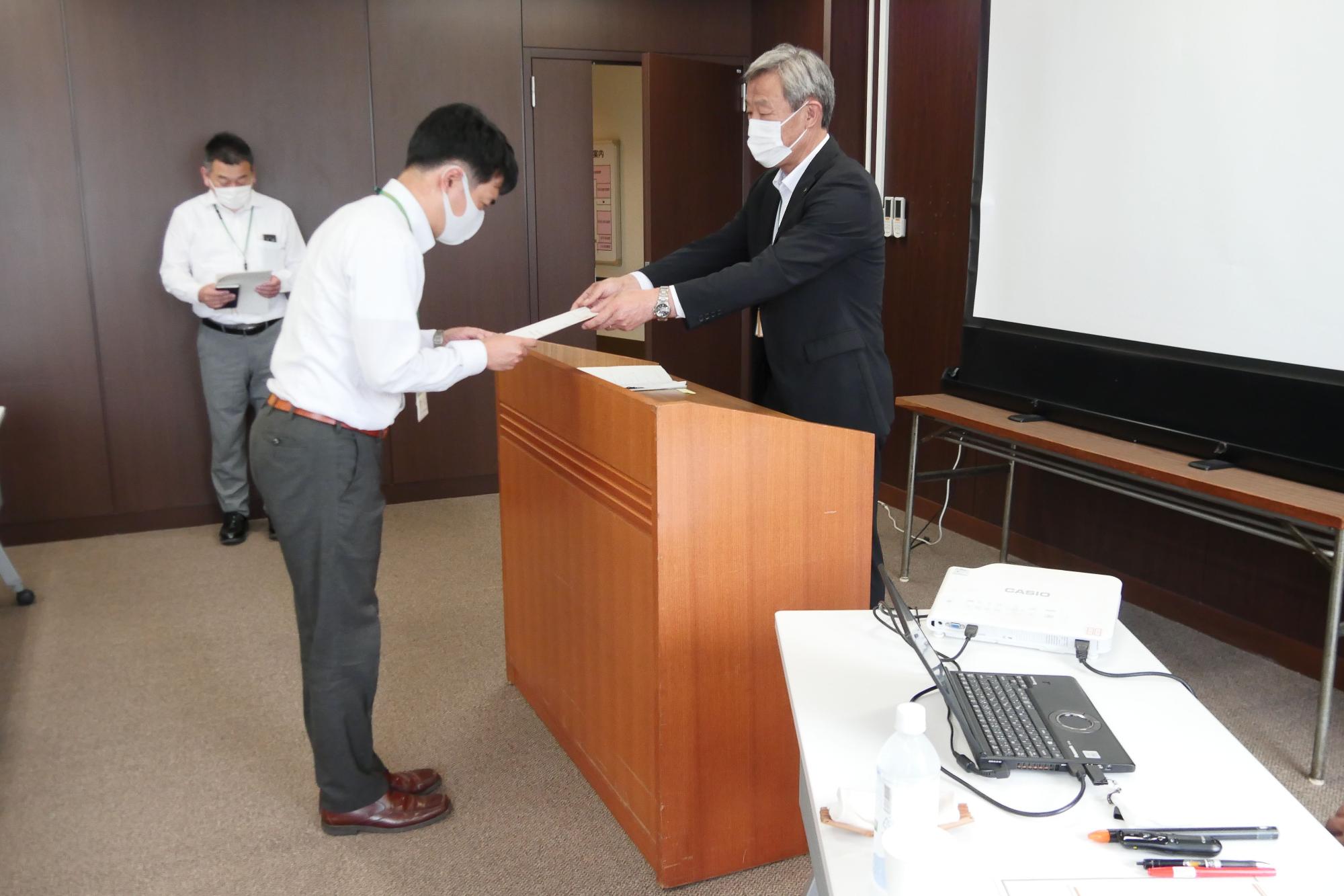 マスクを着用したスーツ姿の男性が、演台越しに、若い男性に書類を手渡している様子の写真