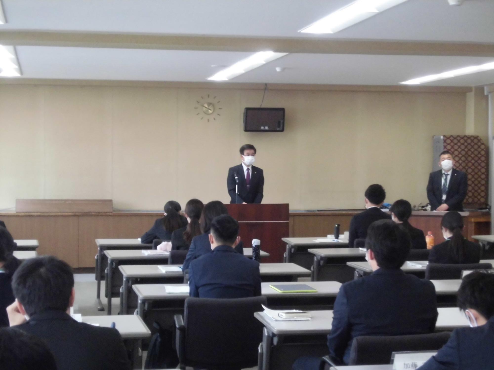 スーツ姿の市長が演台に立って、席に座ったたくさんの参加者たちに向けて話をしている研修会の様子の写真